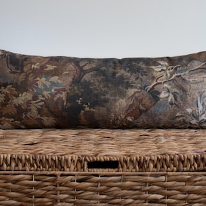 A rectangular decorative pillow with a landscape design featuring trees and foliage in shades of brown, blue, and green. The pillow is placed on top of a woven wicker basket, creating a rustic aesthetic.