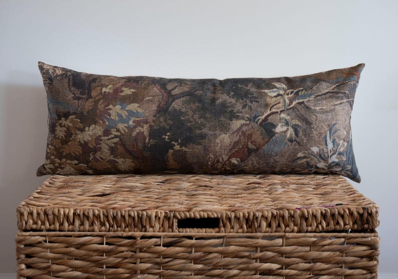 A rectangular decorative pillow with a landscape design featuring trees and foliage in shades of brown, blue, and green. The pillow is placed on top of a woven wicker basket, creating a rustic aesthetic.