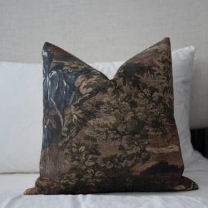 May include: Decorative throw pillow with a detailed tapestry design. The pillow features a landscape scene with trees, foliage, and a muted color palette of brown, green, and blue. The pillow is square and rests on a white surface.