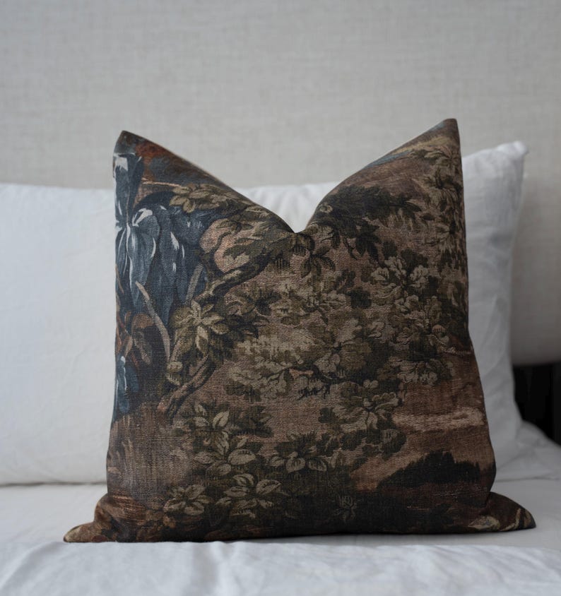 May include: Decorative throw pillow with a detailed tapestry design. The pillow features a landscape scene with trees, foliage, and a muted color palette of brown, green, and blue. The pillow is square and rests on a white surface.