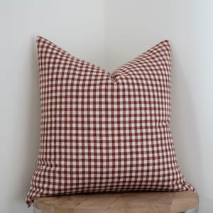 May include: A red and white gingham pillow with a square pattern. The pillow is sitting on a wooden surface.