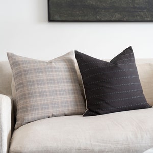 May include: Two throw pillows on a light beige couch. One pillow is a plaid pattern in shades of gray and beige. The other pillow is black with a subtle horizontal stripe pattern.