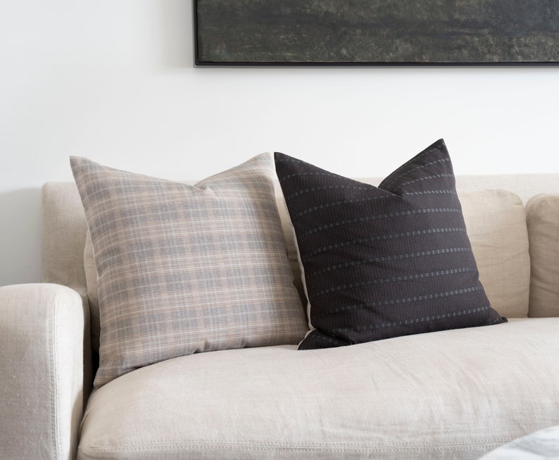May include: Two throw pillows on a light beige couch. One pillow is a plaid pattern in shades of gray and beige. The other pillow is black with a subtle horizontal stripe pattern.