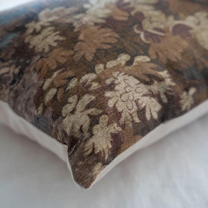 Puede incluir: Coj&iacute;n rectangular decorativo con un estampado floral en tonos marrones, beige y azul. El coj&iacute;n tiene un borde blanco y una textura, lo que sugiere una tela tejida. El dise&ntilde;o presenta hojas y flores estilizadas.