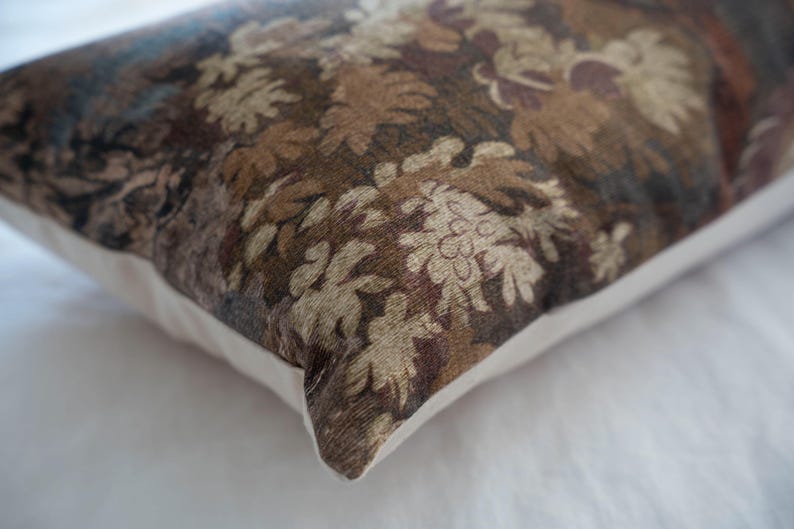 Puede incluir: Coj&iacute;n rectangular decorativo con un estampado floral en tonos marrones, beige y azul. El coj&iacute;n tiene un borde blanco y una textura, lo que sugiere una tela tejida. El dise&ntilde;o presenta hojas y flores estilizadas.