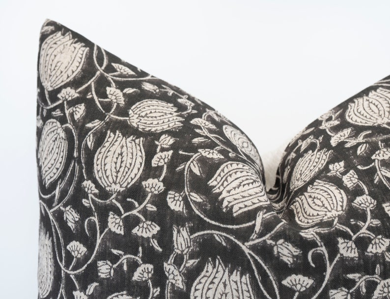 May include: A decorative pillow with a black background and a white floral pattern. The pattern includes stylized flowers and vines. The pillow has a soft, textured appearance and is likely made of fabric. The design is repeated across the pillow's surface.
