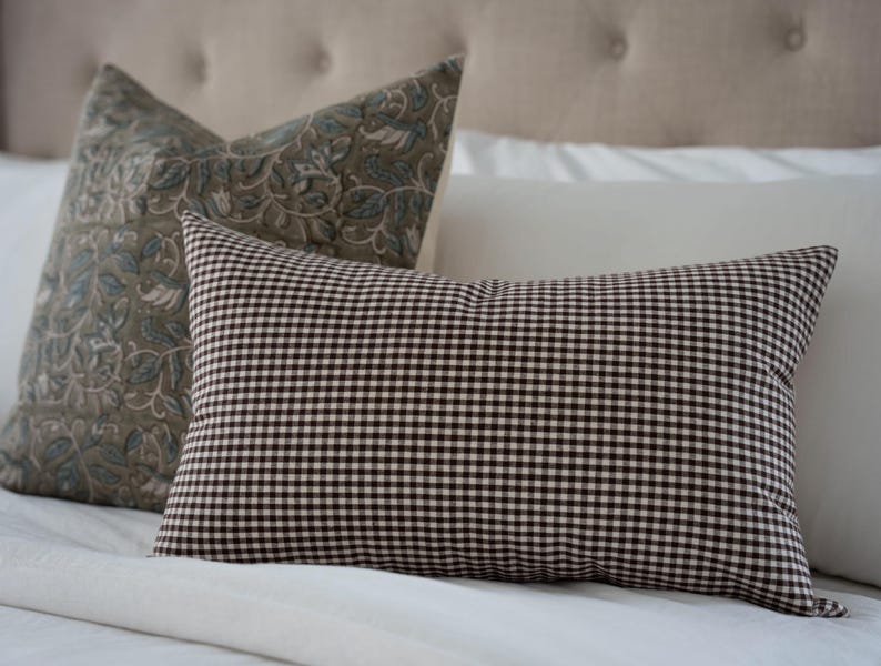K&ouml;nnte beinhalten: Zwei dekorative Kissen auf einem Bett. Ein Kissen hat ein braun-wei&szlig;es Vichy-Muster. Das andere Kissen hat ein Blumenmuster in Gr&uuml;n- und Blaut&ouml;nen. Die Kissen liegen auf einer wei&szlig;en Bettdecke.