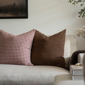 May include: Two throw pillows on a beige couch. One pillow is brown and the other is red and white gingham.