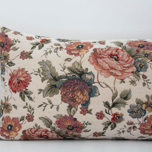 May include: A long rectangular cushion with a floral pattern. The cushion is covered in a cream-coloured fabric with a tapestry-like texture. The floral pattern features red, pink, and green flowers.