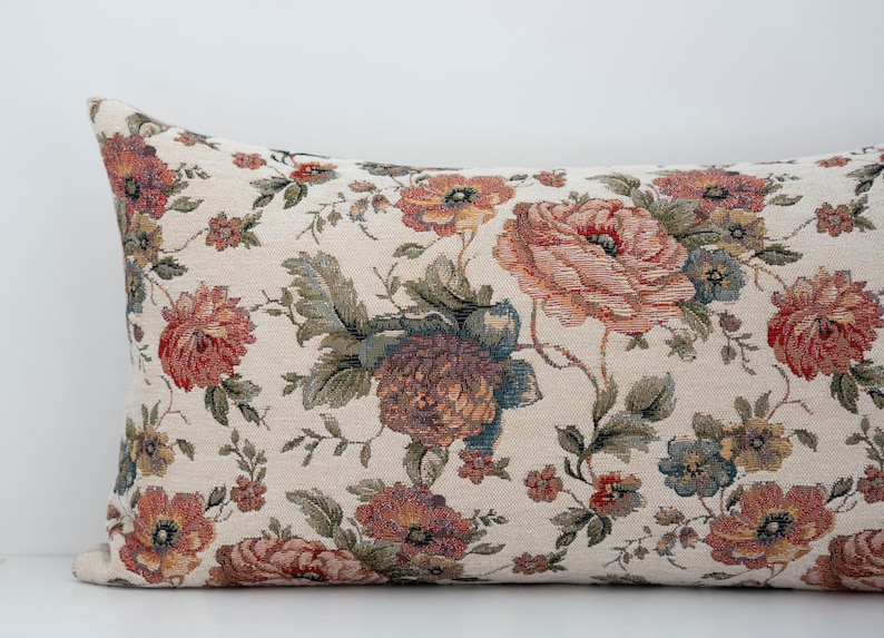 May include: A long rectangular cushion with a floral pattern. The cushion is covered in a cream-coloured fabric with a tapestry-like texture. The floral pattern features red, pink, and green flowers.