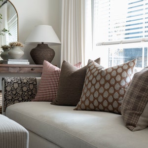 May include: A white sofa with a variety of throw pillows in brown, red, and white patterns. The pillows are arranged in a row on the sofa, with a brown and white patterned throw blanket draped over the back of the sofa.