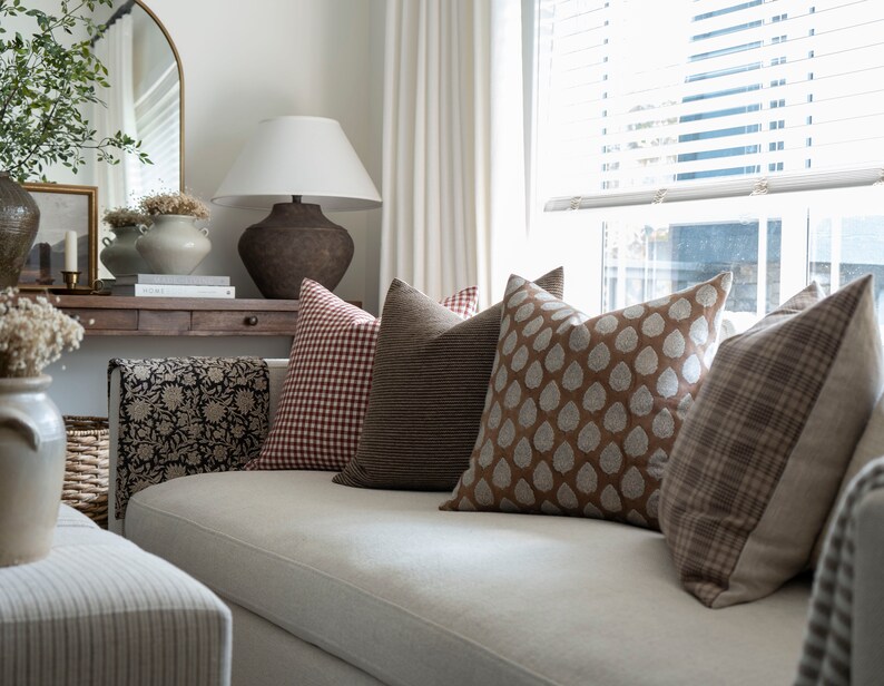 May include: A white sofa with a variety of throw pillows in brown, red, and white patterns. The pillows are arranged in a row on the sofa, with a brown and white patterned throw blanket draped over the back of the sofa.
