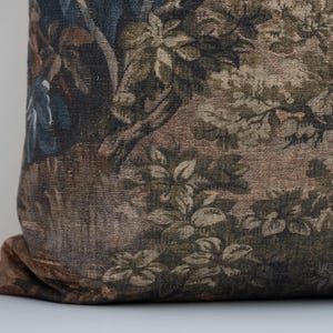 May include: A decorative pillow with a tapestry design. The pillow features a detailed pattern of a forest scene with trees, foliage, and figures in shades of brown, green, and blue. The pillow is resting on a white surface.