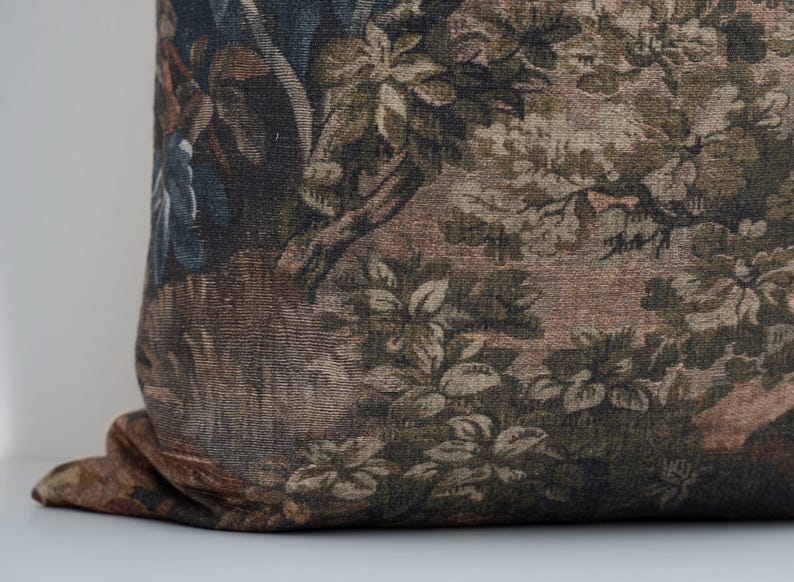 May include: A decorative pillow with a tapestry design. The pillow features a detailed pattern of a forest scene with trees, foliage, and figures in shades of brown, green, and blue. The pillow is resting on a white surface.