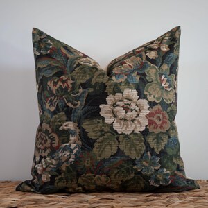 May include: A decorative pillow with a floral pattern in shades of green, blue, and red on a dark background. The pillow features large, stylized flowers and foliage, creating a vintage or traditional aesthetic. The pillow is placed on a woven surface.