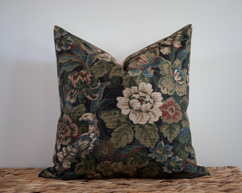 May include: A decorative pillow with a floral pattern in shades of green, blue, and red on a dark background. The pillow features large, stylized flowers and foliage, creating a vintage or traditional aesthetic. The pillow is placed on a woven surface.