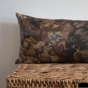 Puede incluir: Un coj&iacute;n rectangular decorativo con un dise&ntilde;o de tapiz que presenta una escena de bosque en tonos marrones, verdes y azules. El coj&iacute;n descansa sobre una cesta de mimbre tejida, creando una est&eacute;tica r&uacute;stica.