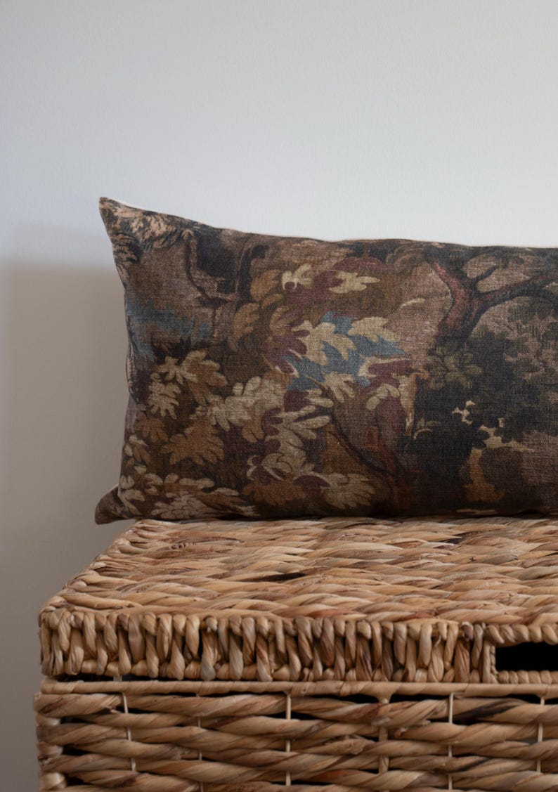 Puede incluir: Un coj&iacute;n rectangular decorativo con un dise&ntilde;o de tapiz que presenta una escena de bosque en tonos marrones, verdes y azules. El coj&iacute;n descansa sobre una cesta de mimbre tejida, creando una est&eacute;tica r&uacute;stica.