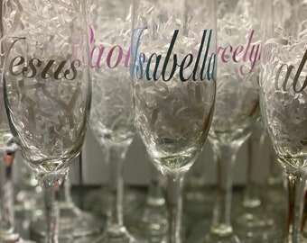 May include: A group of clear glass champagne flutes with personalized names written on them in various colors. The names include "Jesus", "Isabella", "Rebecca", and "Ava".