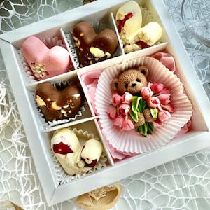 May include: A white box containing assorted chocolates, including heart-shaped ones in pink, brown, and white. A teddy bear with a bouquet of pink flowers is also present in one compartment.