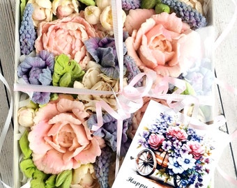 Op de afbeelding: Een witte cadeaudoos gevuld met roze en paarse zeepbloemen. De doos is vastgebonden met een roze lint en een kaart waarop staat "Happy Mother's Day Big Colz! Lauren".