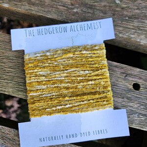 Op de afbeelding: Een spoel met natuurlijk handgeverfde gele en witte vezels, met het label "The Hedgerow Alchemist" en "Naturally Hand Dyed Fibres".