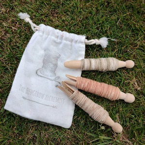 Op de afbeelding: Drie houten spoelen gewikkeld met verschillende kleuren garen, waaronder beige, perzik en groen, in een witte trekkoordtas met de tekst "The Hedgerow Alchemist" erop gedrukt.