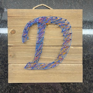 May include: A wooden sign featuring the letter D created using string art. The letter is formed with blue, orange, and purple string wrapped around nails hammered into the wood.