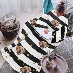 May include: A white blanket with black stripes, gold polka dots, and images of a golden Labrador Retriever. The blanket is draped over a person sitting in a chair, with a book in their lap. The blanket is a cozy home decor item.