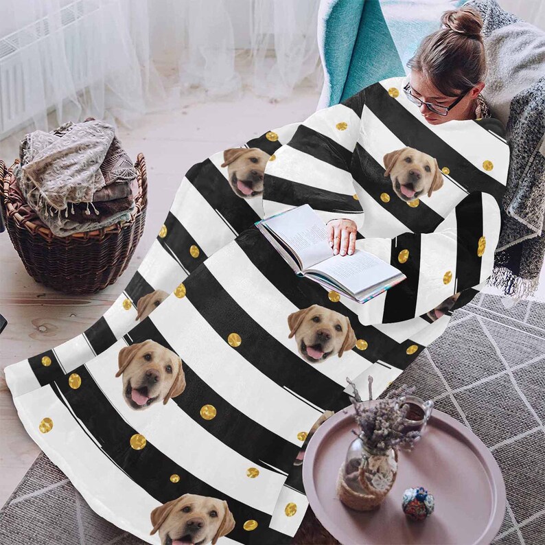 May include: A white blanket with black stripes, gold polka dots, and images of a golden Labrador Retriever. The blanket is draped over a person sitting in a chair, with a book in their lap. The blanket is a cozy home decor item.