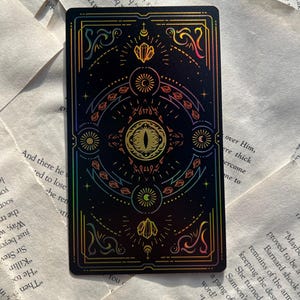 May include: A black tarot card with a holographic design featuring an eye, crystals, and celestial symbols. The card has a colorful, iridescent border and intricate details. The card is rectangular.