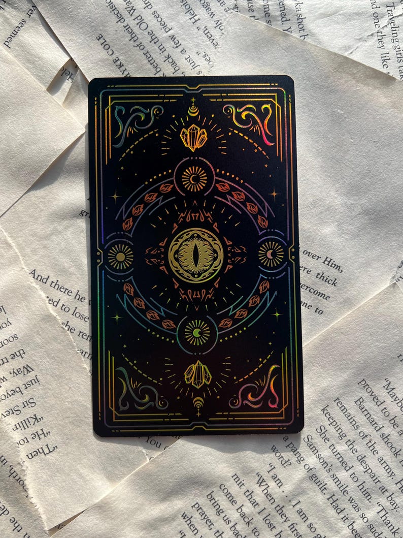 May include: A black tarot card with a holographic design featuring an eye, crystals, and celestial symbols. The card has a colorful, iridescent border and intricate details. The card is rectangular.