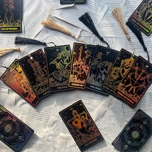 May include: A collection of tarot card bookmarks with gold foil accents and tassel attachments. The cards feature illustrations and titles such as "Two of Swords," "Ace of Cups," and "The Empress." The bookmarks are arranged on a background of open book pages.