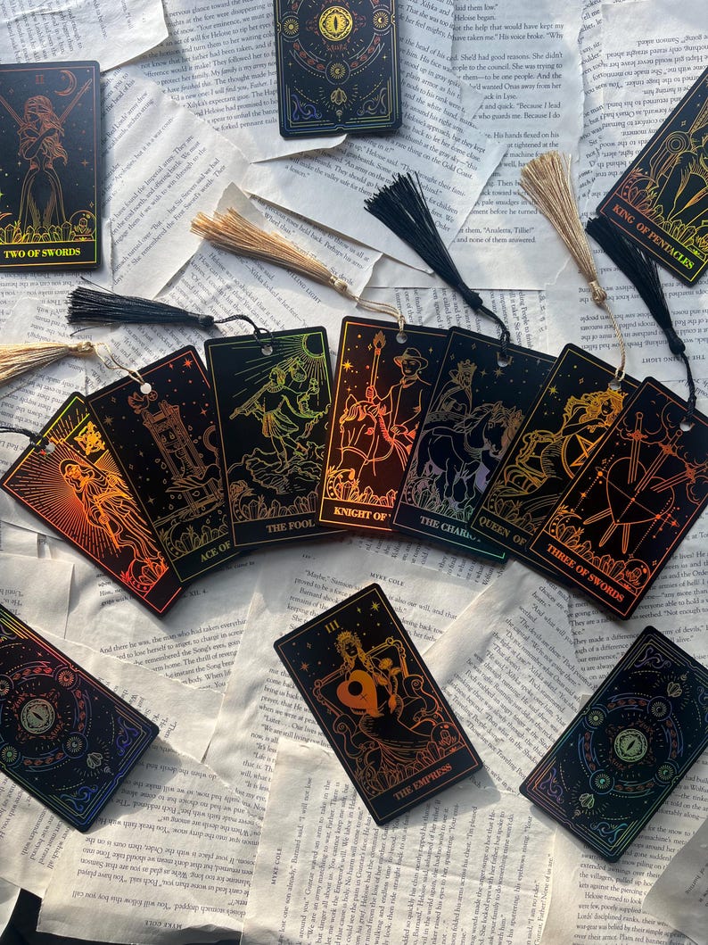 May include: A collection of tarot card bookmarks with gold foil accents and tassel attachments. The cards feature illustrations and titles such as "Two of Swords," "Ace of Cups," and "The Empress." The bookmarks are arranged on a background of open book pages.