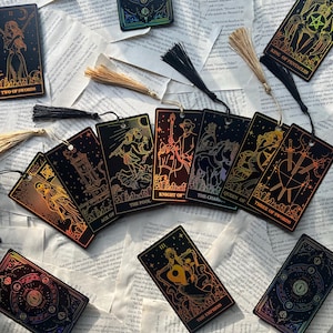 May include: A collection of tarot card bookmarks with gold foil designs and black tassels. The cards feature illustrations and titles like "The Empress," "King of Pentacles," and "Two of Swords." They are arranged on a background of open book pages.
