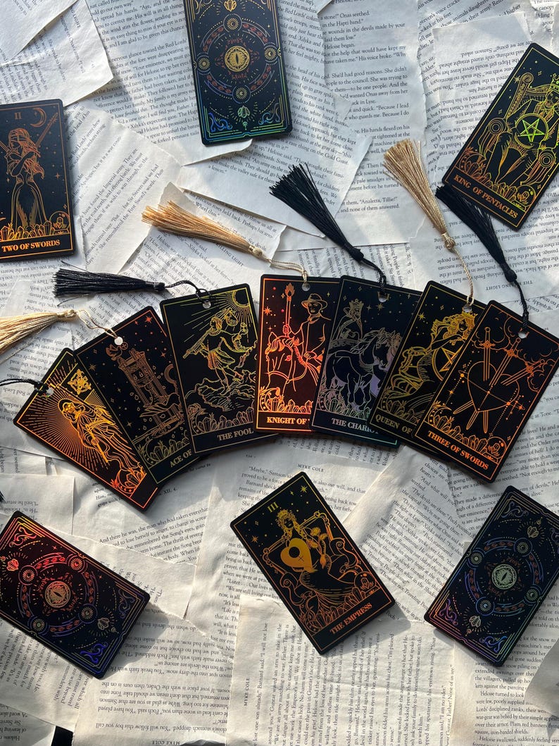 May include: A collection of tarot card bookmarks with gold foil designs and black tassels. The cards feature illustrations and titles like "The Empress," "King of Pentacles," and "Two of Swords." They are arranged on a background of open book pages.