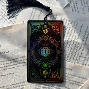May include: Black bookmark with a holographic design featuring celestial and geometric elements, including an eye, crystals, and crescent moons. It has a black tassel. The bookmark is rectangular and rests on open book pages.