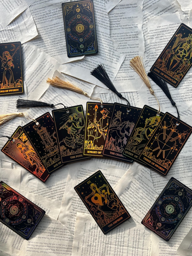 May include: A collection of tarot cards with gold and iridescent foil designs. The cards feature illustrations of figures and symbols, with titles like "Two of Swords," "The Fool," and "The Empress." Each card has a black tassel attached.
