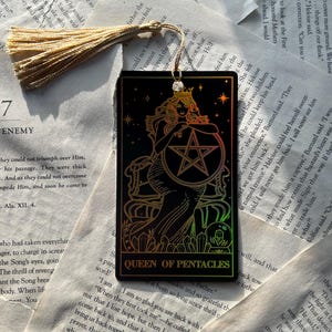 May include: Black tarot card bookmark featuring the Queen of Pentacles. The card has a gold and iridescent design of a woman with a crown, holding a pentacle. A gold tassel hangs from the top.