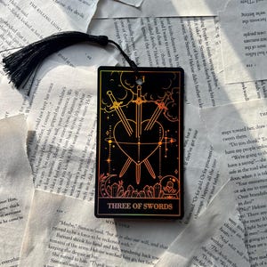 May include: Black bookmark featuring the Three of Swords tarot card design. The card depicts three swords piercing a heart, with a colorful gradient background and the text "THREE OF SWORDS". A black tassel hangs from the top.