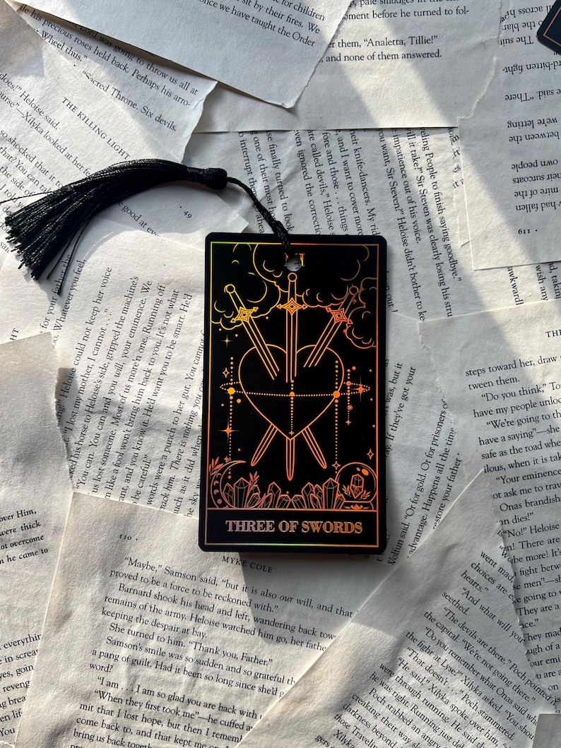 May include: Black bookmark featuring the Three of Swords tarot card design. The card depicts three swords piercing a heart, with a colorful gradient background and the text "THREE OF SWORDS". A black tassel hangs from the top.