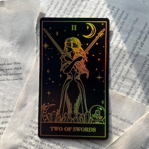 May include: A tarot card featuring a blindfolded figure holding two swords, set against a black background with stars and a crescent moon. The card reads "TWO OF SWORDS" in gold lettering. The card has a holographic, iridescent finish.