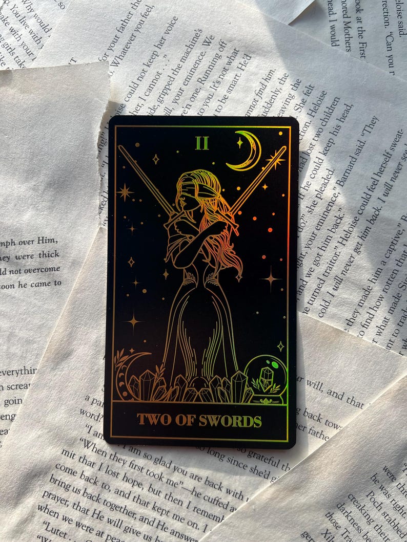 May include: A tarot card featuring a blindfolded figure holding two swords, set against a black background with stars and a crescent moon. The card reads "TWO OF SWORDS" in gold lettering. The card has a holographic, iridescent finish.