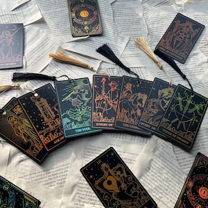 May include: A collection of tarot cards and bookmarks with gold and iridescent accents. The cards feature detailed illustrations and text, including "The Empress" and "King of Pentacles." Tassels are attached to some of the cards, and the items are arranged on a background of open book pages.
