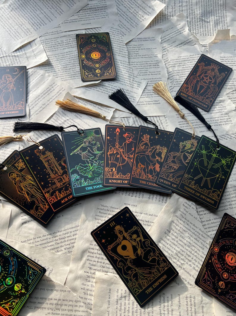 May include: A collection of tarot cards and bookmarks with gold and iridescent accents. The cards feature detailed illustrations and text, including "The Empress" and "King of Pentacles." Tassels are attached to some of the cards, and the items are arranged on a background of open book pages.