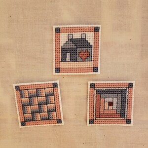 May include: Three small framed cross-stitch patterns. The first pattern is a house with a heart. The second pattern is a quilt block with a checkerboard design. The third pattern is a log cabin quilt block.