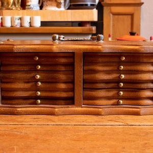 Peut inclure: Un coffret à bijoux en bois vintage avec deux colonnes de tiroirs, chacune avec six petits tiroirs. Les tiroirs ont un design ondulé et sont accentués de petits boutons ronds de couleur dorée. La boîte est en bois aux tons chauds.