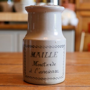 Puede incluir: Un tarro de cerámica color crema con el texto "MAILLE Moutarde à l'ancienne" en escritura negra. El tarro tiene una forma ligeramente acampanada y un borde decorativo alrededor del texto. Está sobre una superficie de madera.