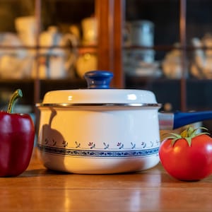 May include: A white enamel saucepan with a blue handle and lid, decorated with a blue floral pattern. A red bell pepper and a red tomato sit beside the pot on a wooden surface. Kitchenware.