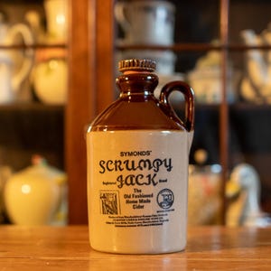 May include: A ceramic jug with a brown top and handle, and a cream-colored body. The jug has the text "Symonds Scrumpy Jack" and "The Old Fashioned Home Made Cider" printed on it. The jug is sitting on a wooden surface.